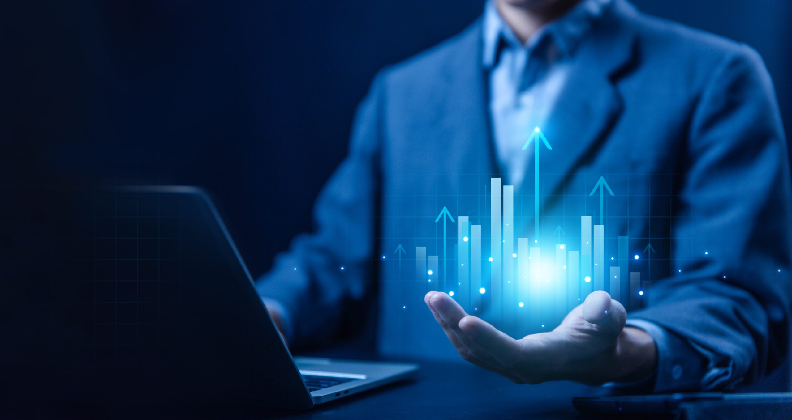 A person in a blue suit sits at a desk with a laptop, holding a glowing, upward-trending bar graph and arrows above their open palm. The digital chart suggests growth and positive trends in a business or financial context, illuminated against a dark background.