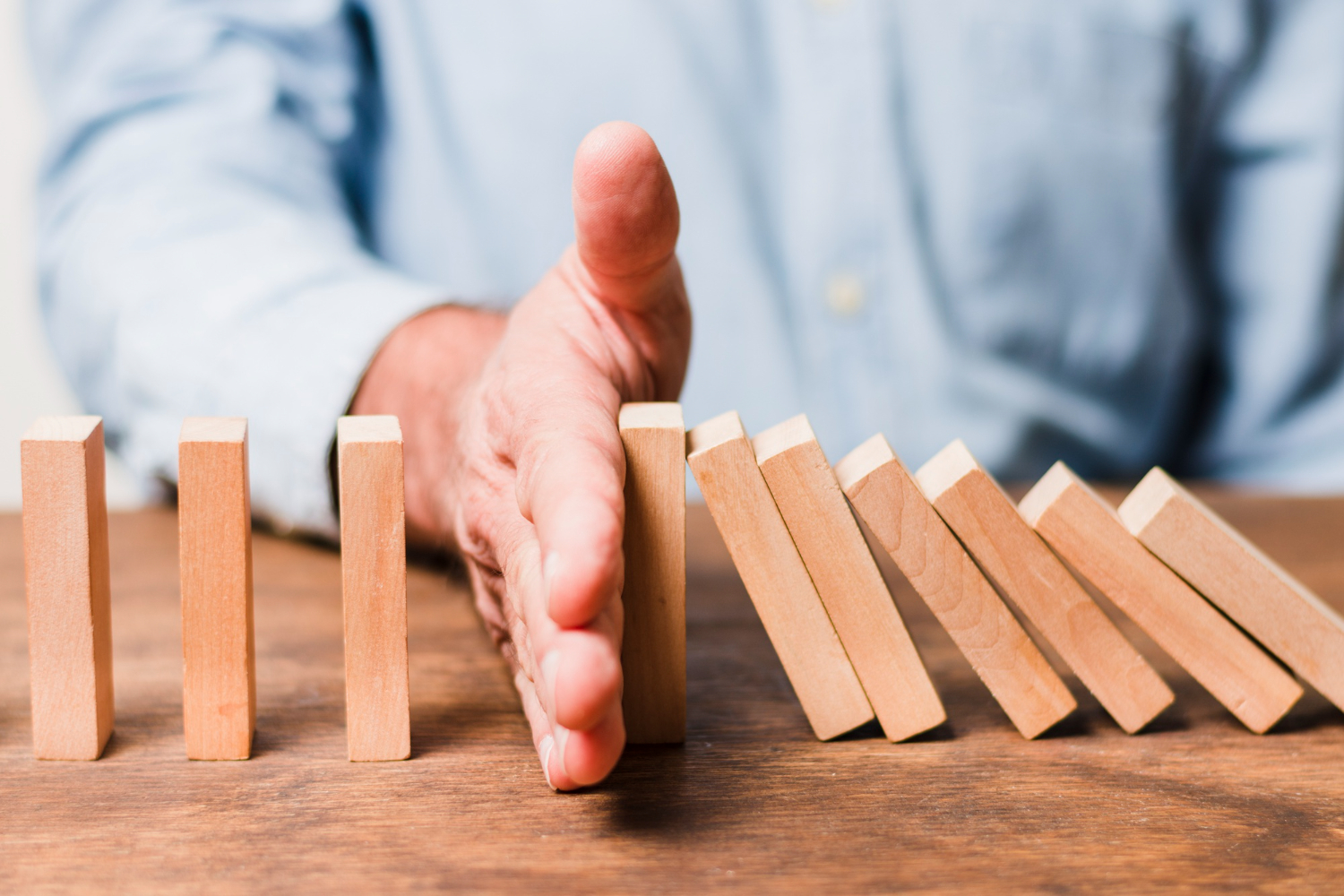 Hand stopping a row of wooden domino blocks from falling, preventing a chain reaction.