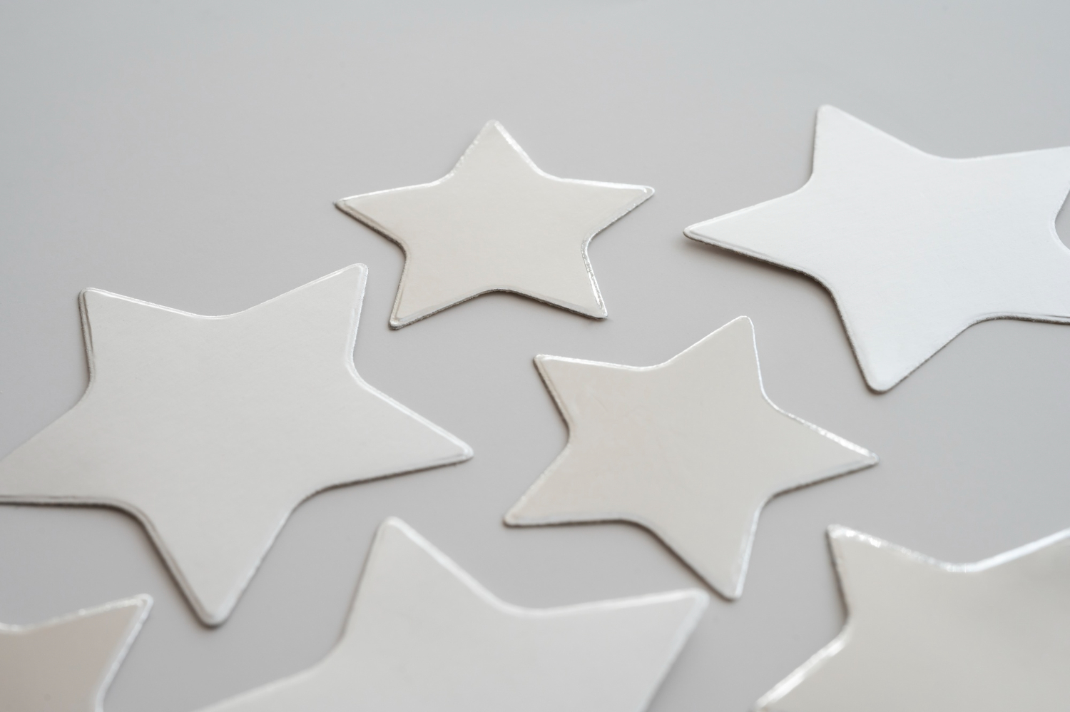 A top-down, minimalist view of several silver or light-gray star-shaped cutouts scattered across a flat, neutral-colored surface. The lighting is soft, creating subtle shadows and highlights on the shapes.