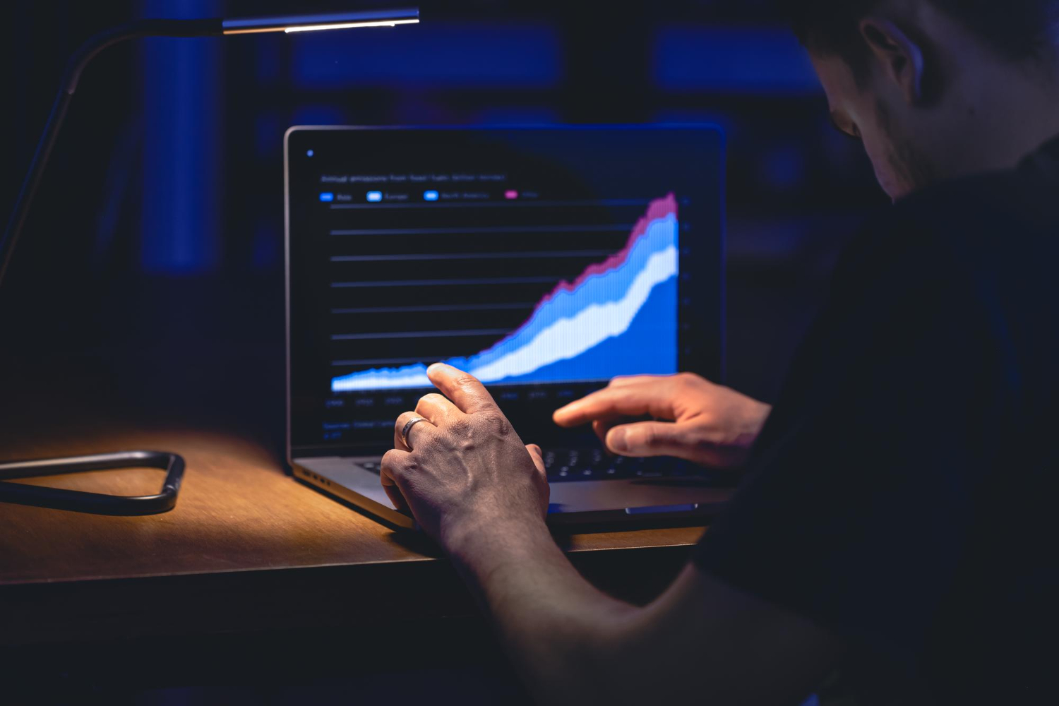 An over-the-shoulder view of a person working at a laptop in a dimly lit room at night. The screen displays a colorful area chart showing a strong upward growth trend. A small desk lamp illuminates the workspace.